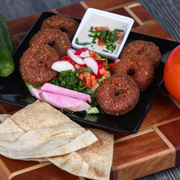 A black plate of Lebanese falafel, salad, pickled turnips, tahini, and pita bread on a wooden board.