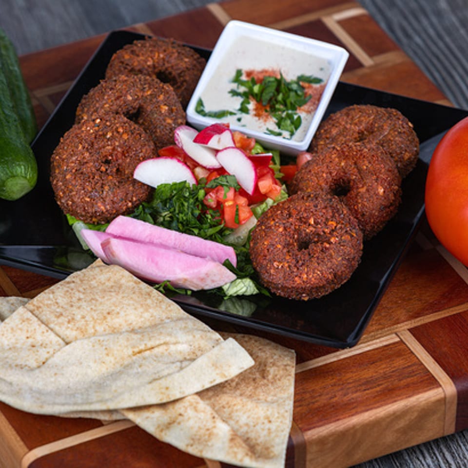 A black plate of Lebanese falafel, salad, pickled turnips, tahini, and pita bread on a wooden board.