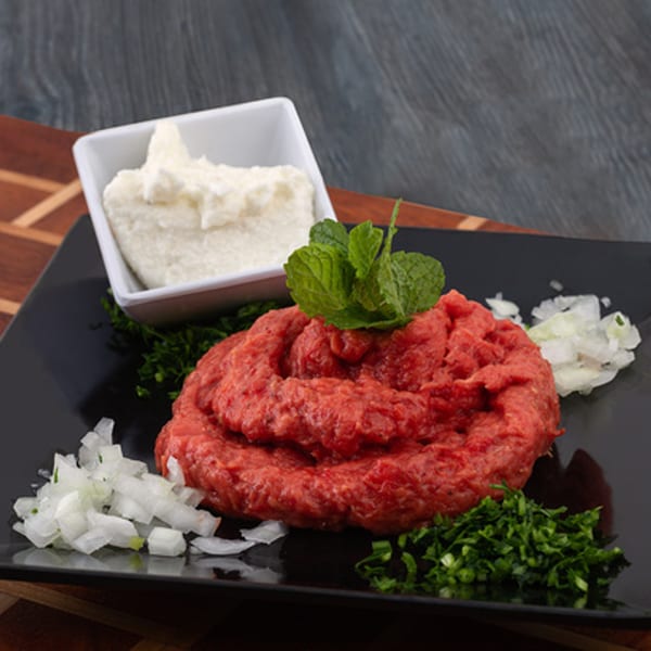 Raw kibbeh with mint, chopped onions, parsley, and a side of white dip on a black plate.