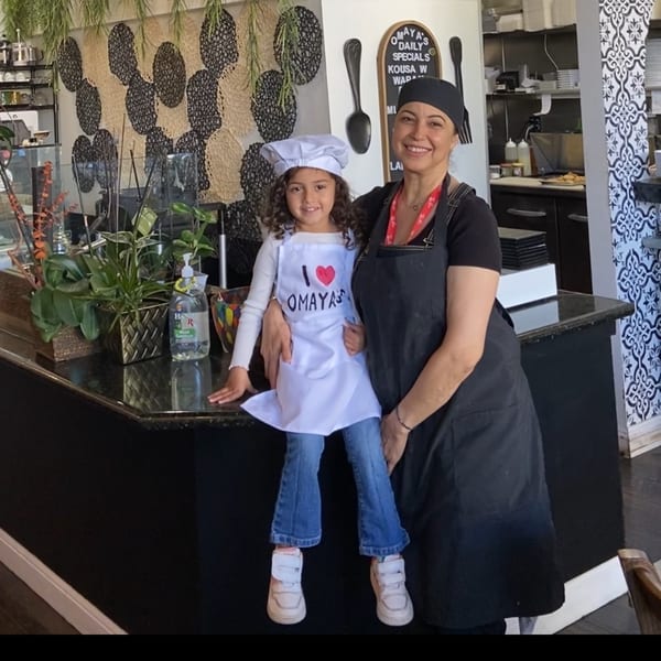 A woman and a young girl in chef attire smiling behind the counter at Omaya's Lebanese Cuisine.