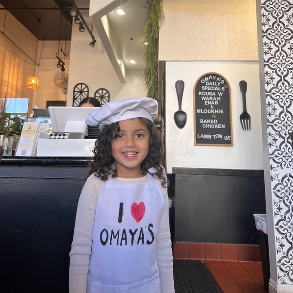 A smiling child in a chef's hat and "I love Omaya's" apron stands in Omaya's Lebanese Cuisine.