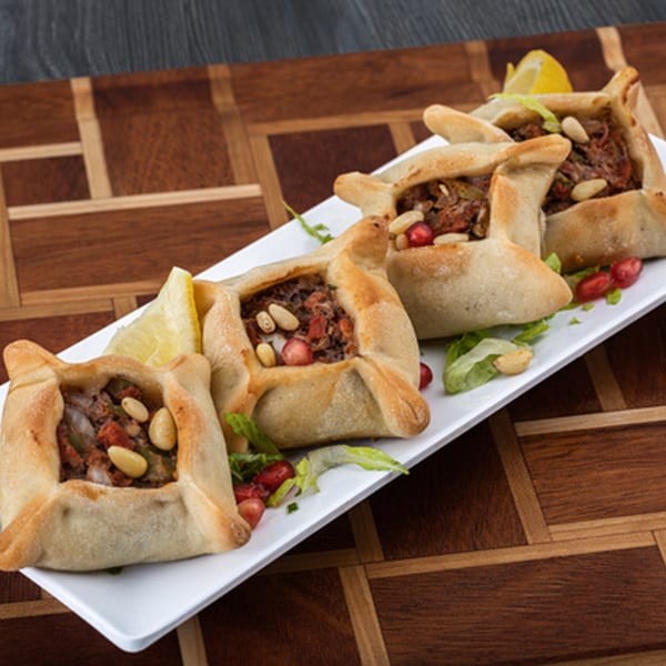 Four savory Lebanese meat pies, fatayer, garnished with pine nuts and pomegranate seeds on a white plate.