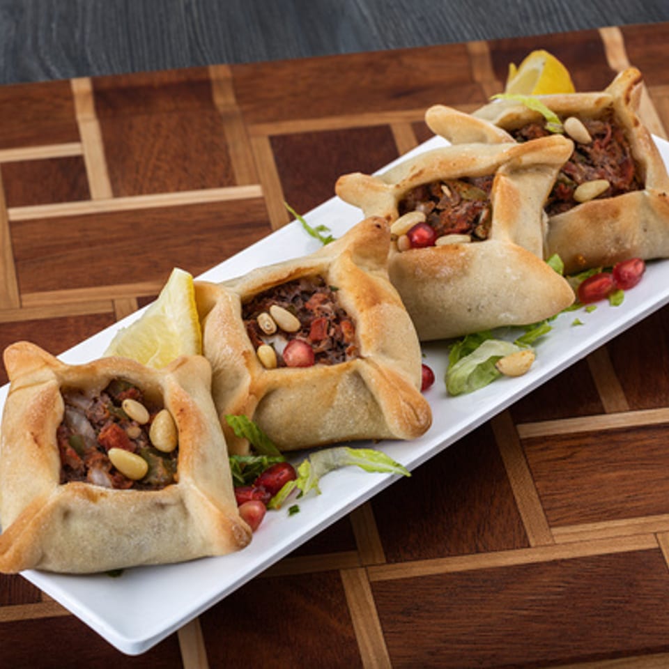 Four savory Lebanese meat pies, fatayer, garnished with pine nuts and pomegranate seeds on a white plate.