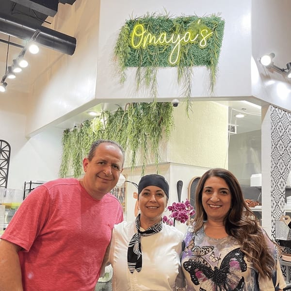 Three smiling people, two women and one man, standing in front of Omaya's Lebanese Cuisine sign.