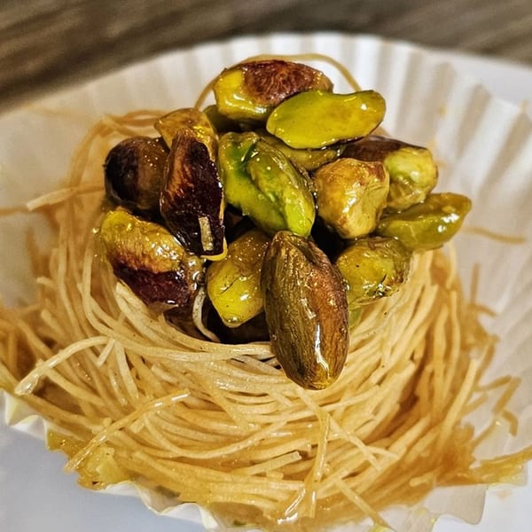 Lebanese bird's nest baklava with shredded phyllo dough and glistening green pistachios in a white paper cup.